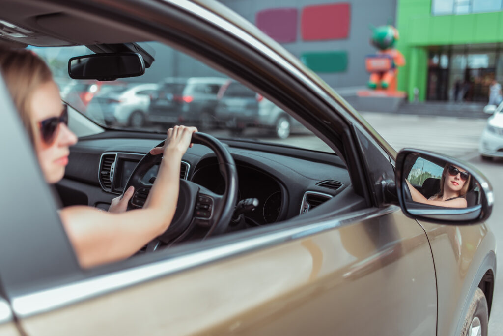 A girl driving a right-hand drive car is parked in a parking lot, near a shopping center