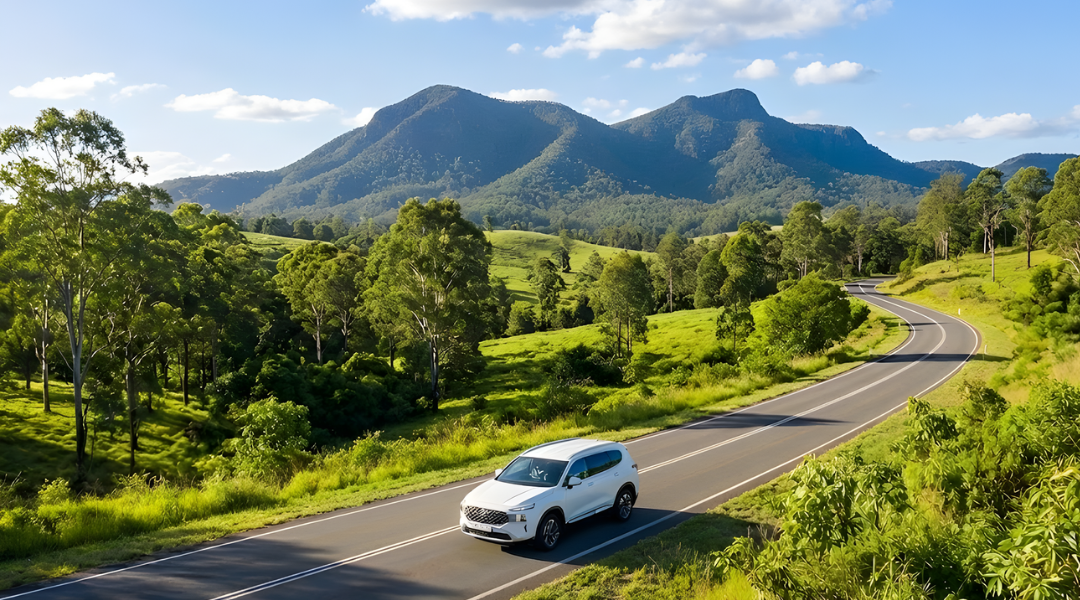 white SUV driving on a sealed country road with lush green mountains in the background (evoking the Scenic Rim or Glass House Mountains). Bright, sunny Queensland weather.