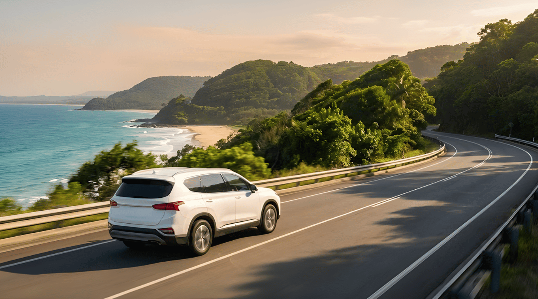 Intermediate SUV driving along a coastal highway with lush greenery (Northern NSW vibe). The ocean is visible in the background.