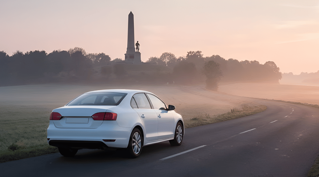 a clean, white, unbranded sedan driving on a quiet country road at sunrise. In the background, a silhouette of a classic Australian war memorial (obelisk or soldier statue) is visible on a hill.