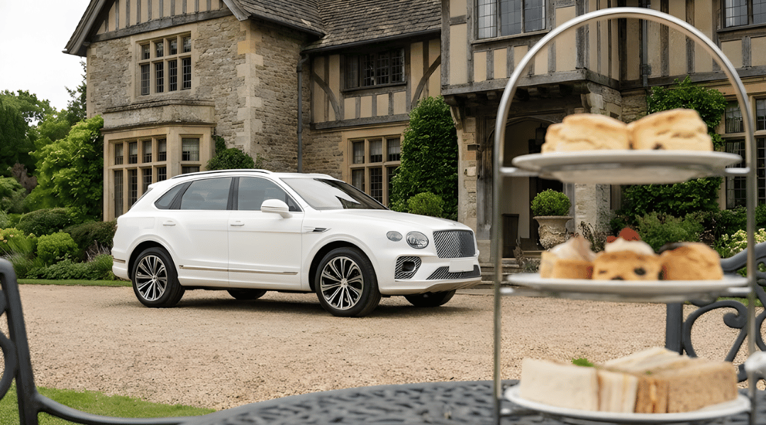 luxury SUV parked on a driveway in front of a heritage-style country estate or garden. In the foreground (soft focus), a tiered high tea stand with scones and sandwiches is visible on a garden table.