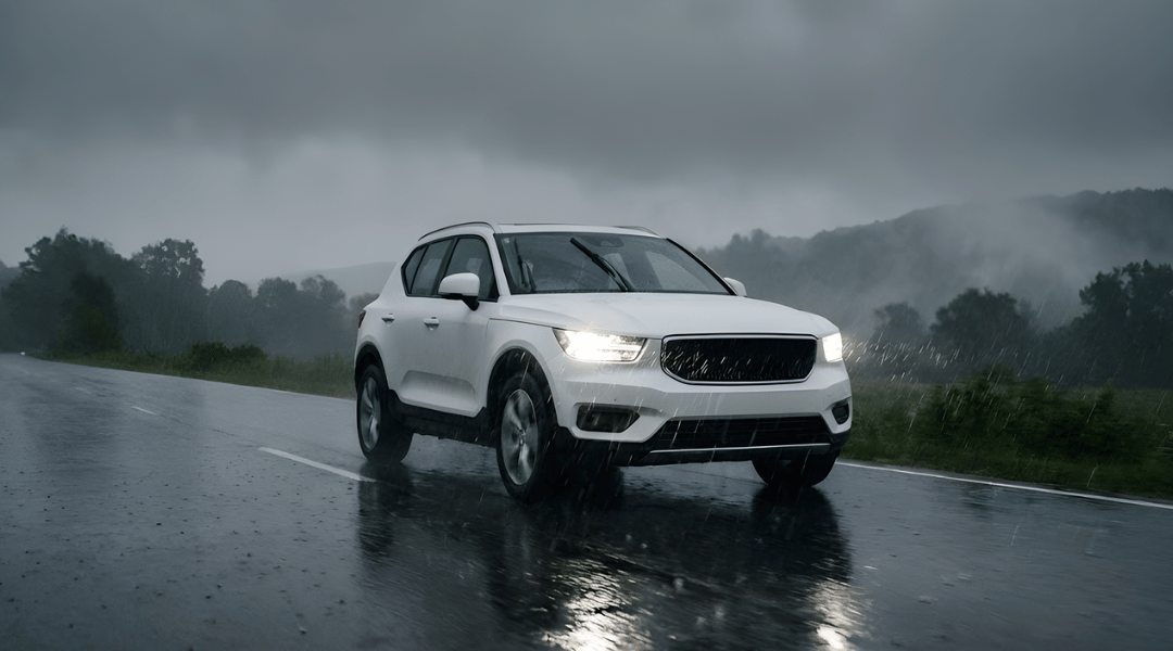 SUV driving on a wet asphalt road during a rainy day. The car's headlights are on, reflecting on the wet surface. Raindrops are visible in the air
