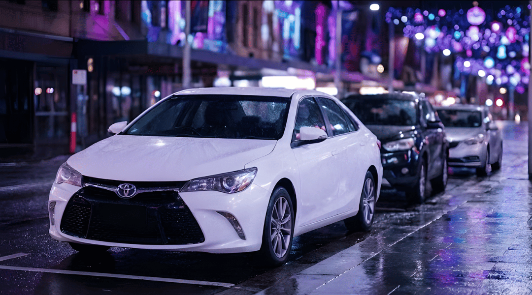 sedan driving on a wet city street at night. The background is filled with vibrant, colourful out-of-focus city lights
