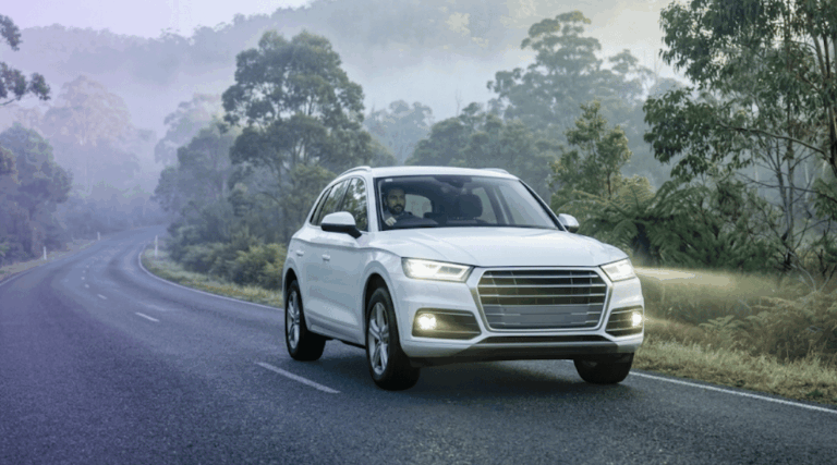 A white rental SUV safely navigating a misty Australian country road with its headlights on during an early morning drive.
