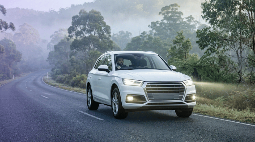 A white rental SUV safely navigating a misty Australian country road with its headlights on during an early morning drive.