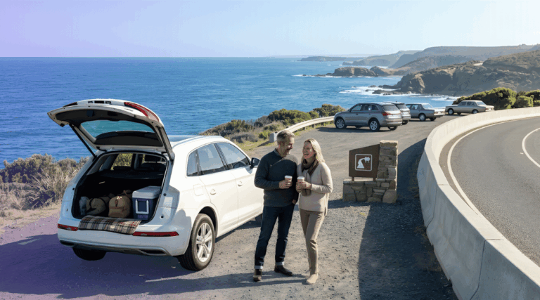 A mature couple enjoying a morning coffee beside their white luxury SUV on a scenic Australian coastal road.