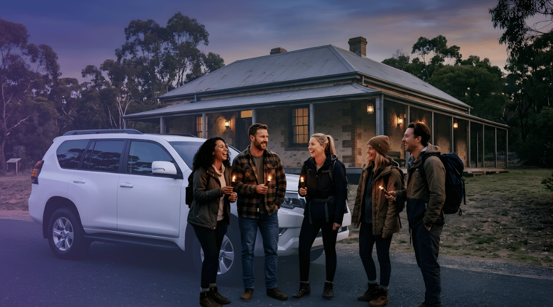 A group of friends laughing beside their white rental SUV as they arrive at a historic Australian building for a Halloween ghost tour.