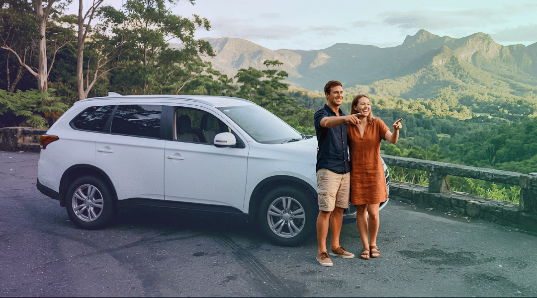 A smiling couple admiring the lush rainforest views next to their white rental SUV in the Gold Coast hinterland.