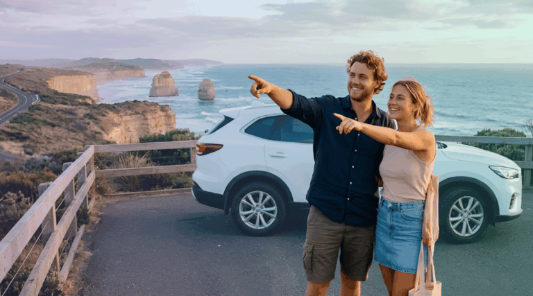 A smiling couple admiring the spectacular coastal views next to their white rental SUV along the Great Ocean Road.