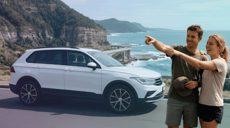 A smiling couple admiring the iconic Sea Cliff Bridge next to their white rental SUV on the Grand Pacific Drive.