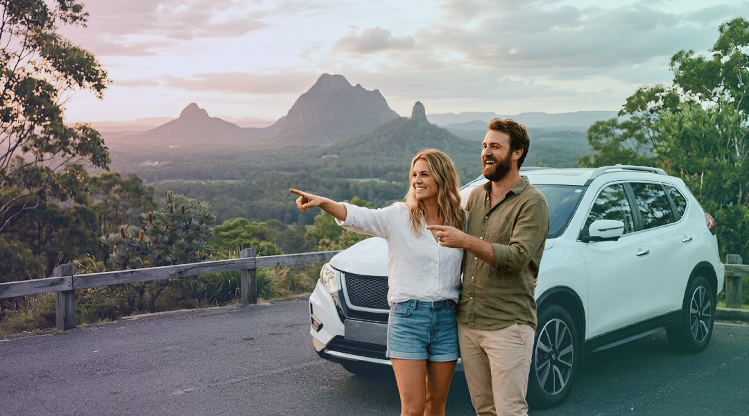 A smiling couple admiring the Glass House Mountains next to their white rental SUV while bypassing highway traffic.
