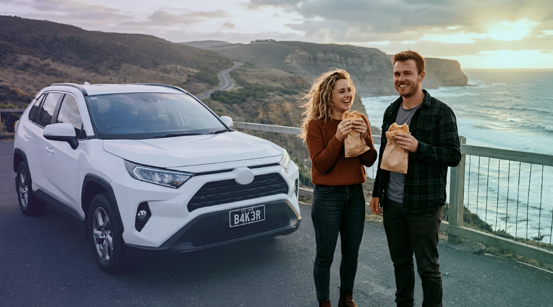 A smiling couple eating meat pies next to their white rental SUV while enjoying a coastal Victorian road trip.