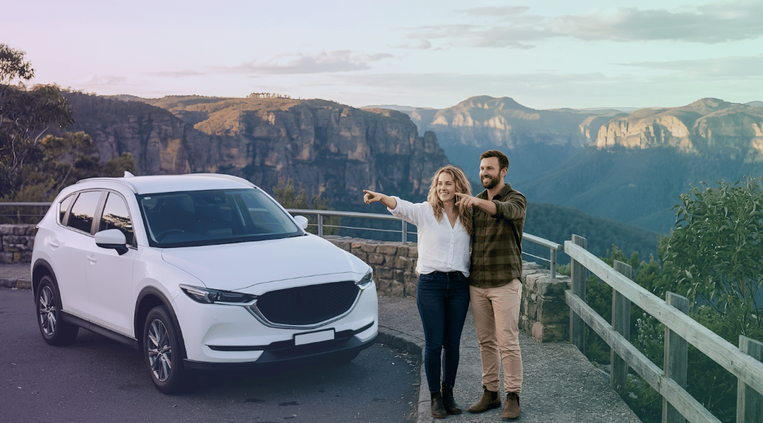 A smiling couple admiring the vast Blue Mountains valleys next to their white rental SUV at a quiet, secluded lookout.
