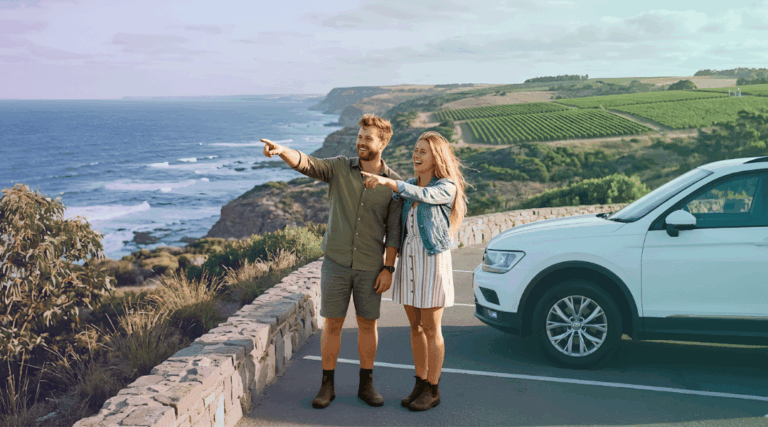 A smiling couple next to their white rental SUV deciding between the coast and the vineyards near Melbourne.