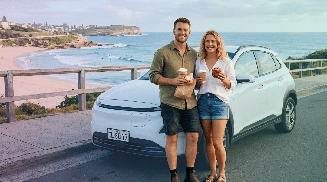 A smiling couple holding takeaway coffees and pastries next to their white rental SUV while enjoying a Sydney coastal morning.