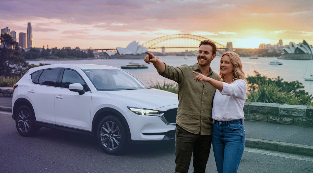 A smiling couple admiring a quiet Sydney Harbour sunset next to their white rental SUV.